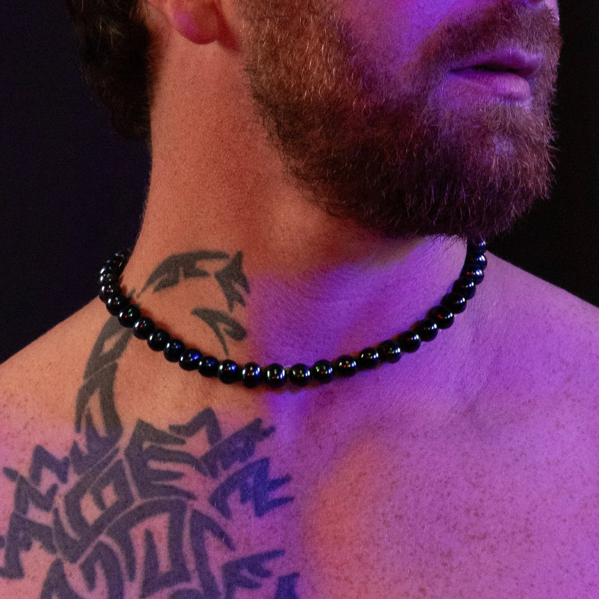 Black beaded necklace worn against tattooed skin under purple lighting.