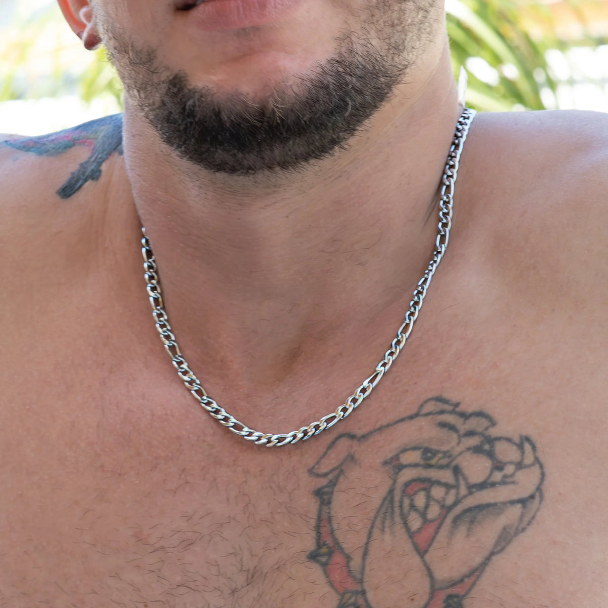 Silver chain necklace worn around a person’s neck with visible tattoos and facial hair.