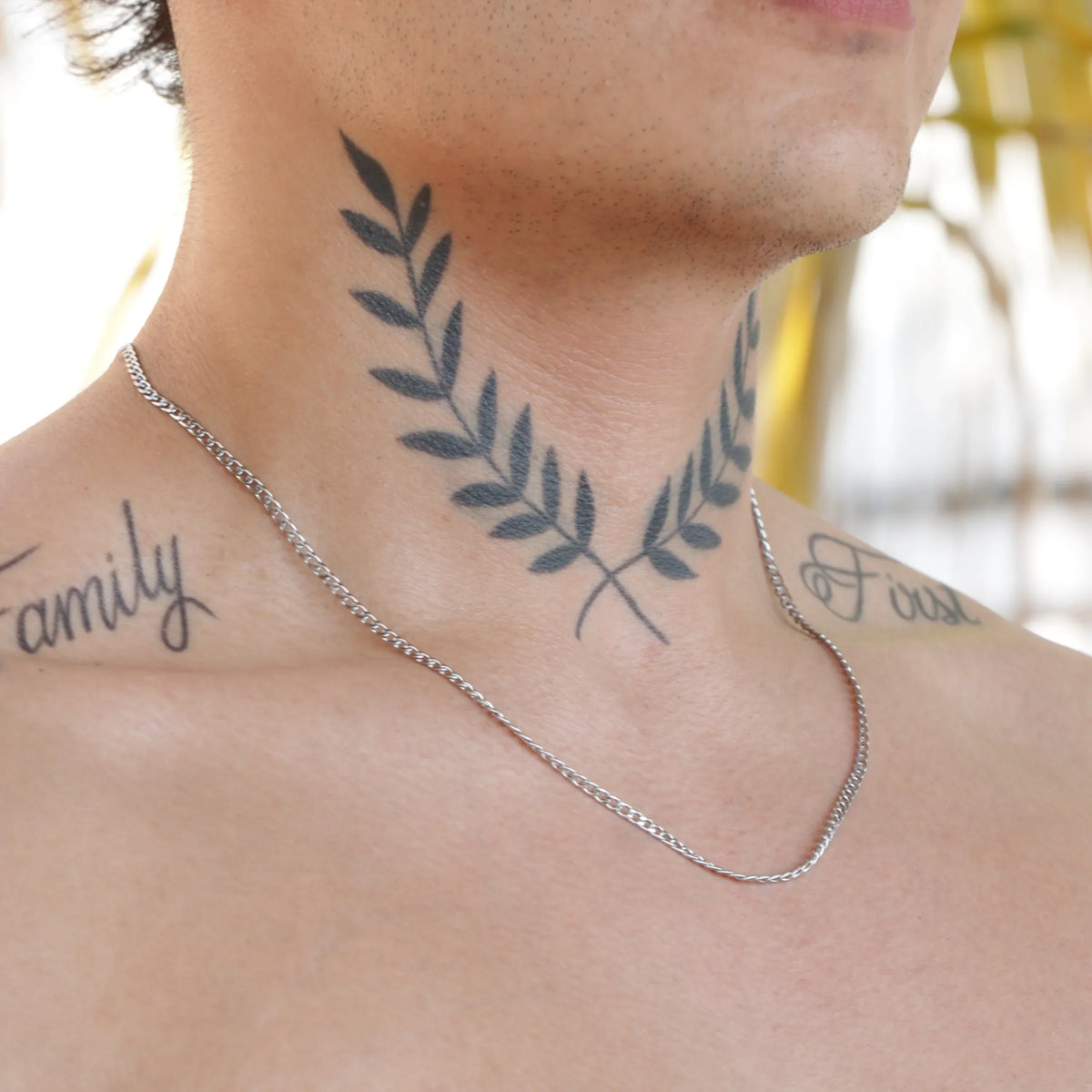 Neck tattoo featuring symmetrical olive branches in blue-green ink with ’family’ text visible on the side.