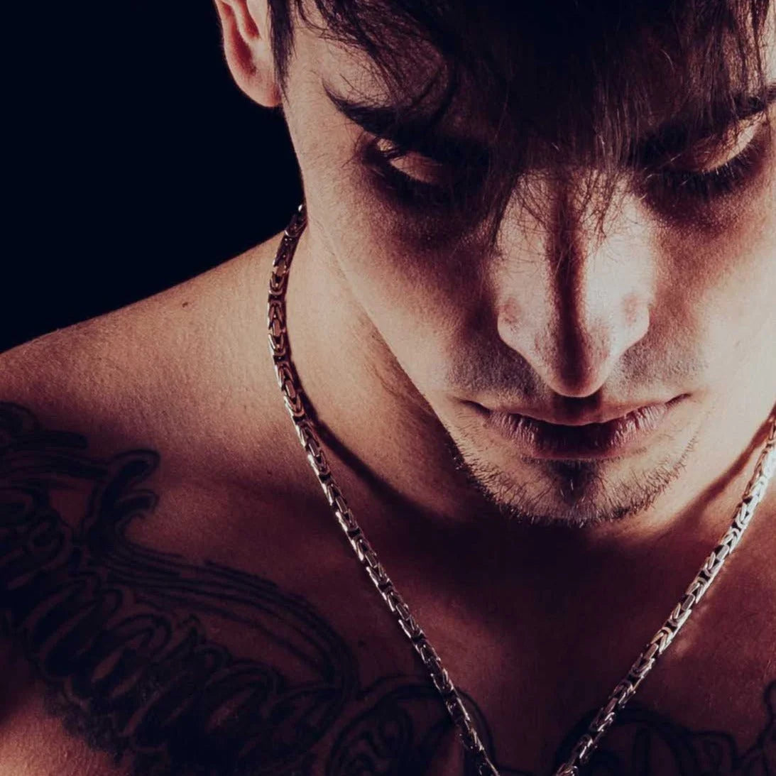 Close-up portrait of a man with dark hair, facial hair, and visible tattoos on his shoulder, wearing a chain necklace.