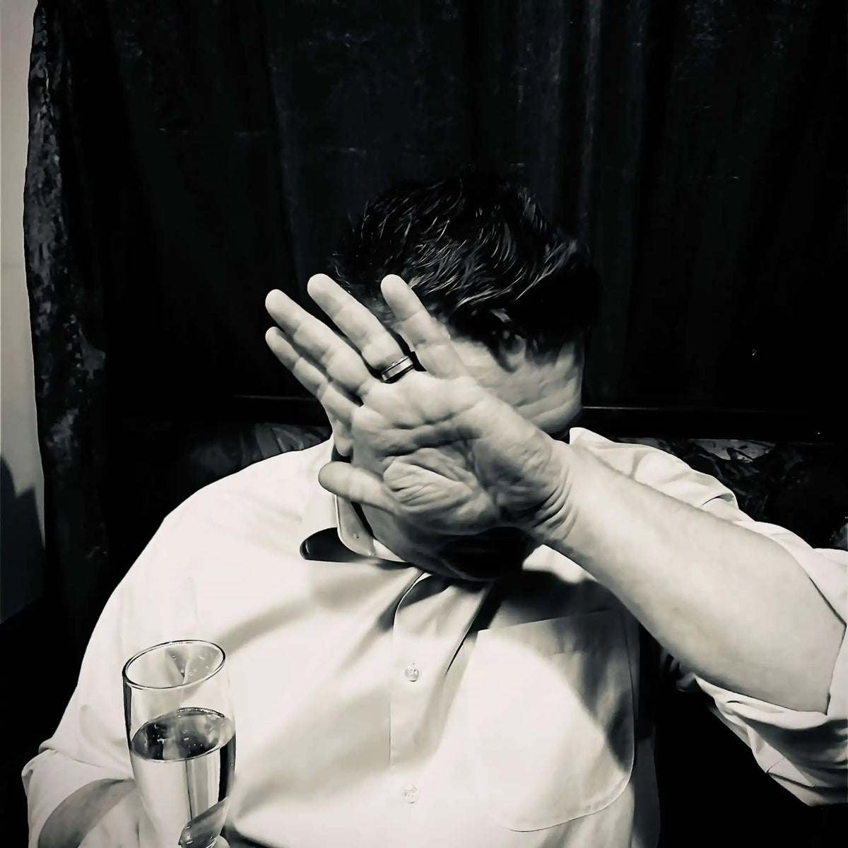 A person covering their face with both hands while sitting at a table with a glass of water.