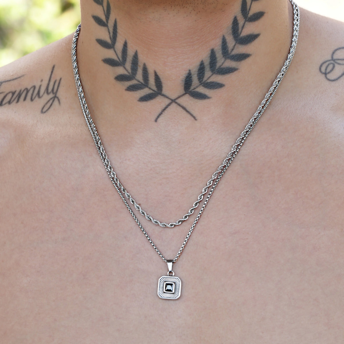 Necklace with a square pendant and a laurel wreath tattoo visible on the wearer’s neck.