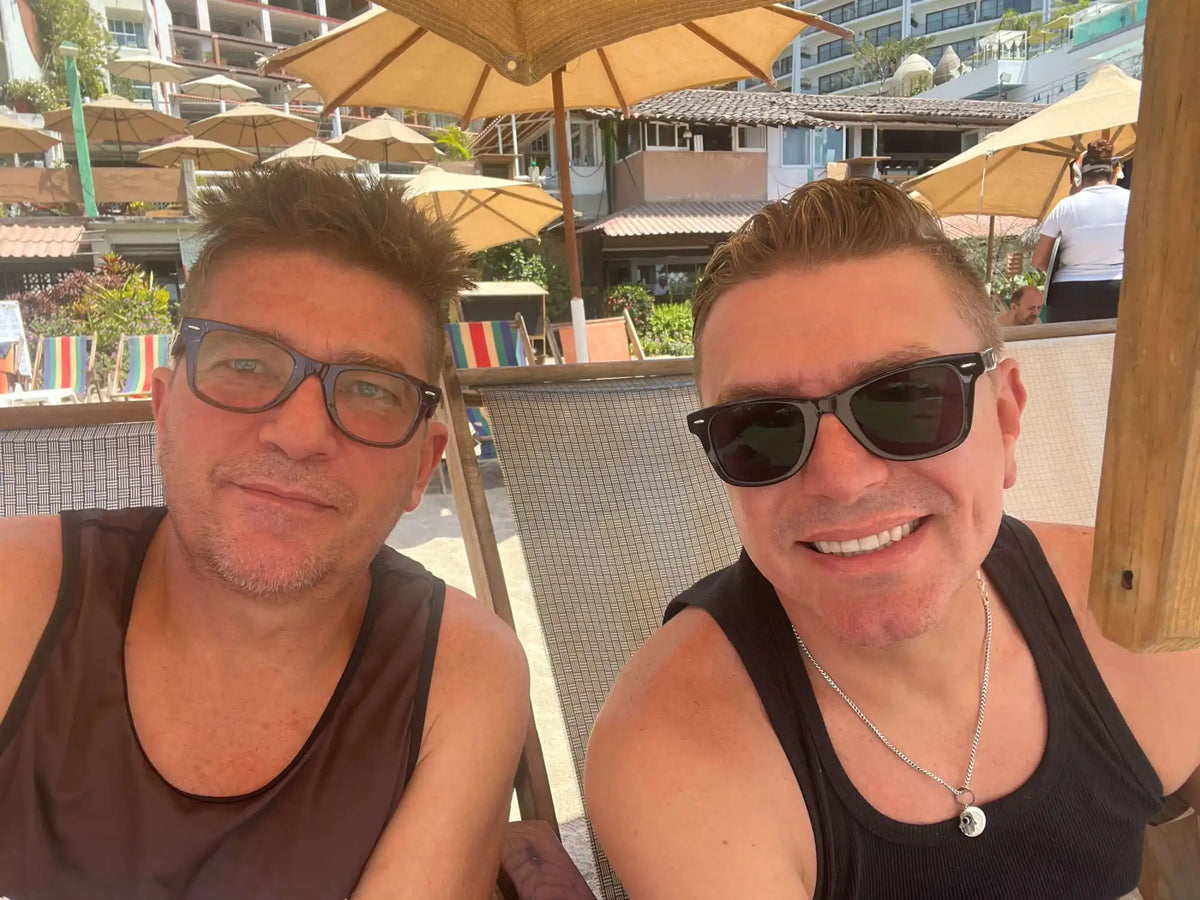 A selfie of two people wearing tank tops and sunglasses relaxing under patio umbrellas.