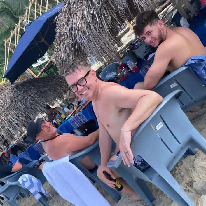 Three shirtless people relaxing on beach chairs under a thatched umbrella.